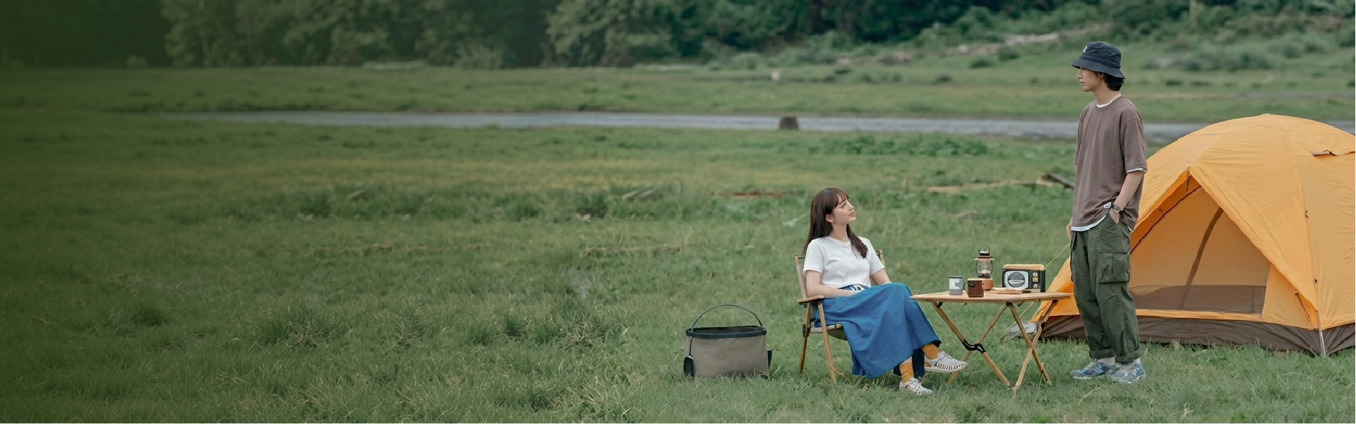 A wide outdoor lifestyle shot of three campers relaxing at a grassy campsite next to an orange tent. A woman in a mustard yellow bucket hat sits in a folding chair by a wooden table, while a man in a black Yama Workwear Bucket Hat stands nearby wearing an earth-toned outfit. The scene highlights rugged outdoor style and utility gear framed by an open field and natural greenery at Grandmama Store in Hong Kong.