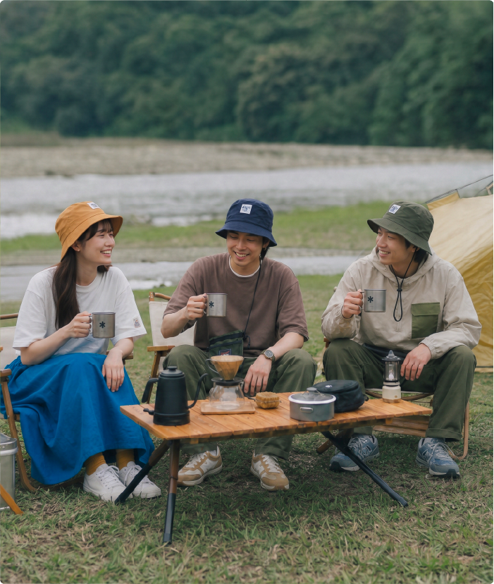 A full lifestyle group photo featuring three models outdoor camping in the Yama Series style. A woman sits in a folding chair wearing a mustard yellow bucket hat, a man seated beside her wears a dark navy blue Yama Workwear Bucket Hat while sipping coffee, and a third man stands in an olive green bucket hat and beige jacket near their tent at Grandmama Store in Hong Kong.