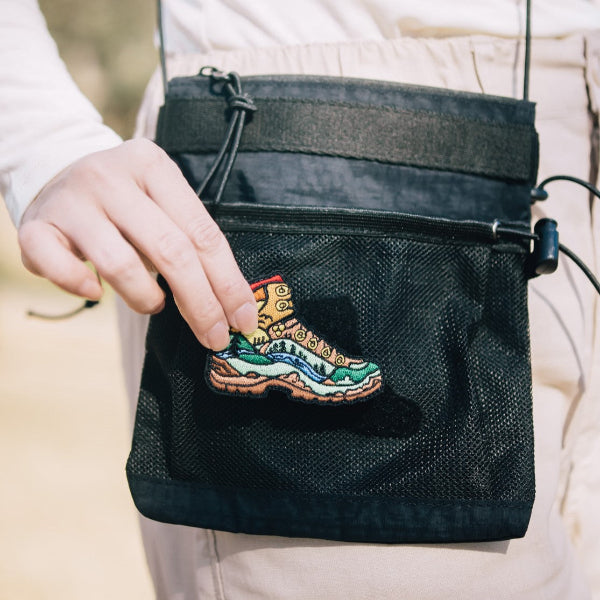 Hand applying an embroidered hiking boot patch to the front mesh pocket of the Yama Multifunctional Bag｜Black at Grandmama Store in Hong Kong.