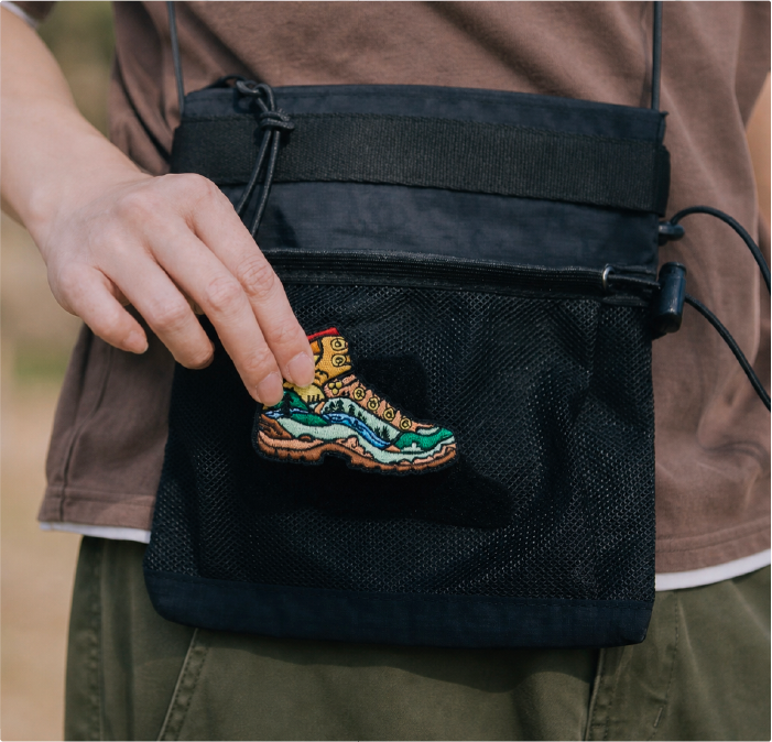 A close-up view of a person attaching an embroidered hiking boot patch to a black mesh front pocket of a sacoche crossbody bag, styled as part of the Yama Series workwear outfit. The image highlights the durable nylon fabric, cord cinches, and modular utility design of the outdoor accessories line at Grandmama Store in Hong Kong.