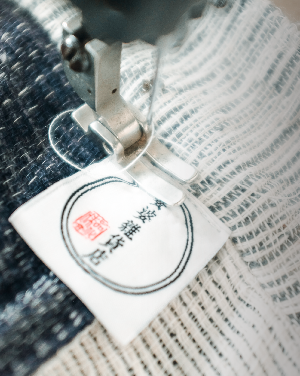 A close-up of a silver sewing machine presser foot stitching a white fabric brand label, which features a circular logo with traditional Chinese characters and a red seal stamp, onto a textured indigo and white woven fabric at Grandmama Store in Hong Kong