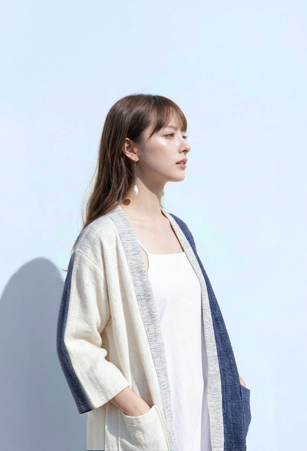 A woman with long brown hair in profile view wearing a hand-cranked Kimono cardigan in Mt. Fuji color—featuring a cream-white body, light gray textured trim, and a deep indigo blue back and sleeve accent—styled over a simple white dress, captured against a clean, light blue background at Grandmama Store in Hong Kong.