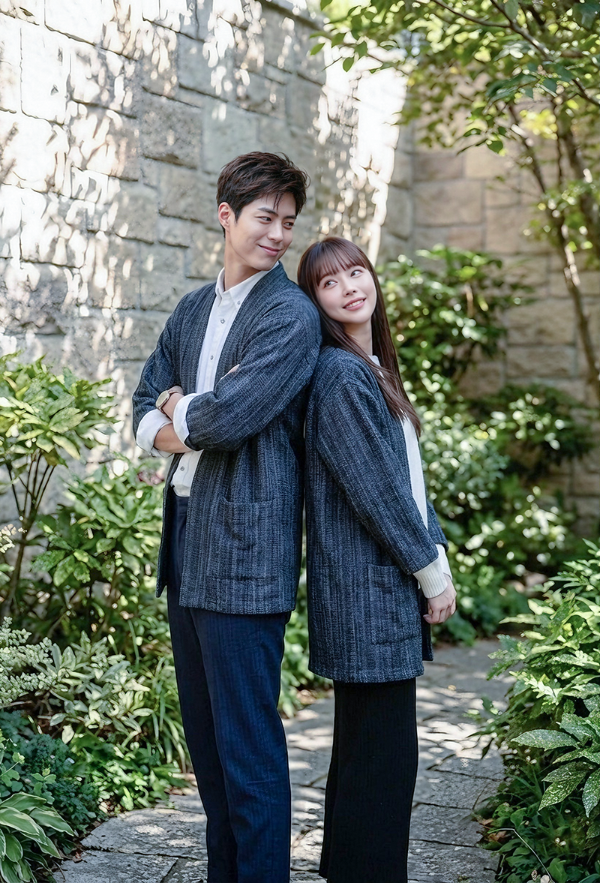A smiling man and woman standing back-to-back on a stone path wearing matching unisex Hand-cranked Kimono cardigans in Miho Matsubara color—a deep charcoal gray or faded black handwoven textured fabric with subtle vertical grain detailing and front pockets—against a rustic stone wall background surrounded by lush green foliage at Grandmama Store in Hong Kong.