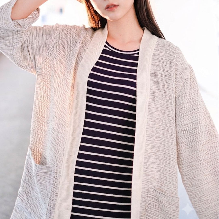A close-up shot of a model wearing an open-front, hand-cranked Kimono cardigan in the Aurora Snow colorway—featuring a highly textured cream and light beige slub-knit handwoven cotton fabric, a matching solid cream collar trim, and front patch pockets—styled over a navy and white striped shirt at Grandmama Store in Hong Kong.