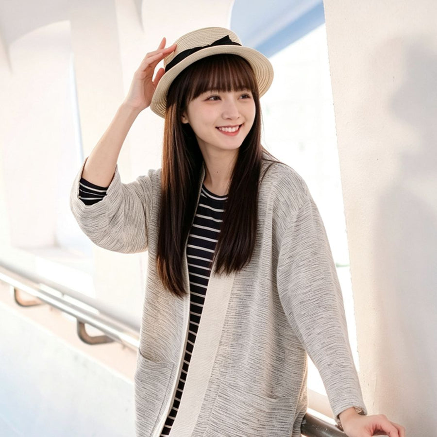 A smiling young woman with long brown hair and bangs wearing a straw fedora hat, standing outdoors next to a railing while wearing a hand-cranked Kimono cardigan in the Aurora Snow colorway—featuring a heavily textured cream and light gray slub-knit handwoven cotton fabric, wide cuffs, and a solid cream trim—styled open over a navy and white striped top at Grandmama Store in Hong Kong.