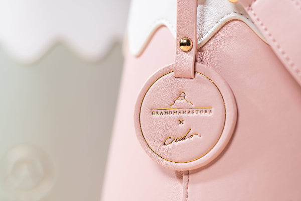 A close-up view focusing on the pink-colored panel and matching circular leather charm of the heavily textured leather Mt. Fuji Goen Bag, referenced as image_1.png. The photograph highlights the distinct slub-knit handwoven cotton texture of the bag's exterior. A precise, embossed circular logo featuring Mt. Fuji and the gold-stamped collaboration text, 'GRANDMAMA STORE X Crudo,' is centrally positioned on the pink tag at Grandmama Store in Hong Kong.