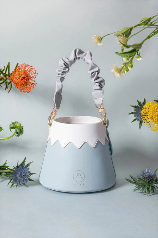 A front flat lay view of the Mt. Fuji Goen Bag in the 'Snow' colourway, centered on a light-blue background and surrounded by floating wildflowers. The image displays the bag’s smooth, premium leather texture in its signature split-panel design, featuring a wavy white snow-cap top and a pastel light-blue base. It showcases the circular embossed mountain logo, gold hardware, and a unique, ruffled scrunchie-style light-blue leather shoulder strap at Grandmama Store in Hong Kong.
