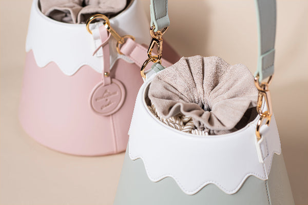 A close-up studio shot featuring two Mt. Fuji Goen Bags arranged together: one in a soft pastel pink colourway in the background and one in a muted sage-like forest green colourway in the foreground. The image highlights the distinctive split-panel design with a wavy white top mimicking Mt. Fuji's snow cap, showcasing the smooth, premium texture of the leather panels, gold hardware clasps, and a soft beige fabric drawstring interior pouch closure at Grandmama Store in Hong Kong.